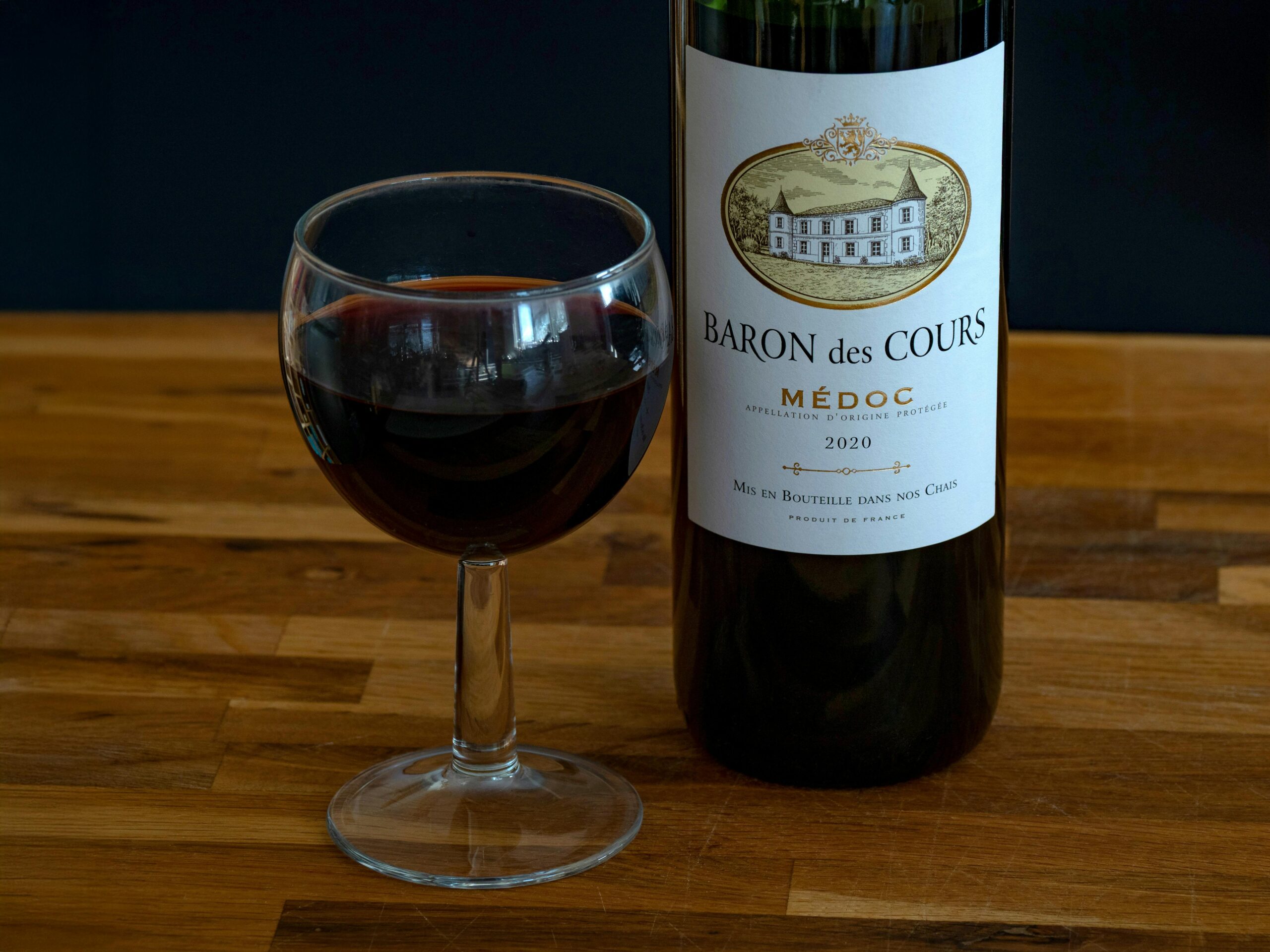 Carte Close-up shot of a Baron des Cours 2020 Médoc wine bottle with a filled glass on a wooden table.